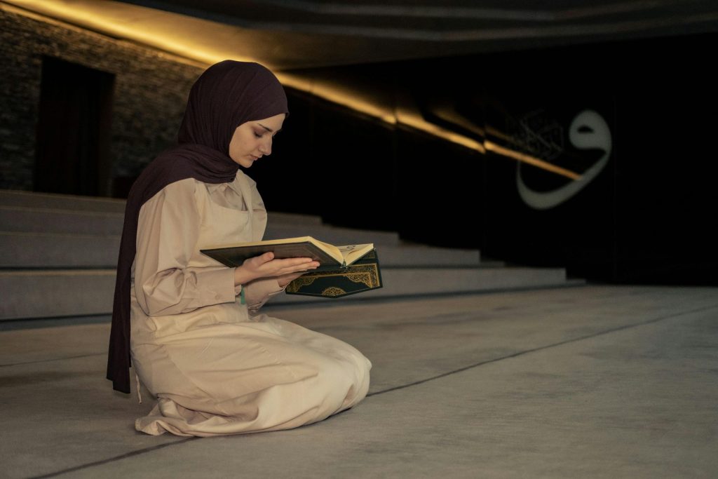 A woman kneeling and reading a Koran inside a dimly lit mosque, wearing a hijab.