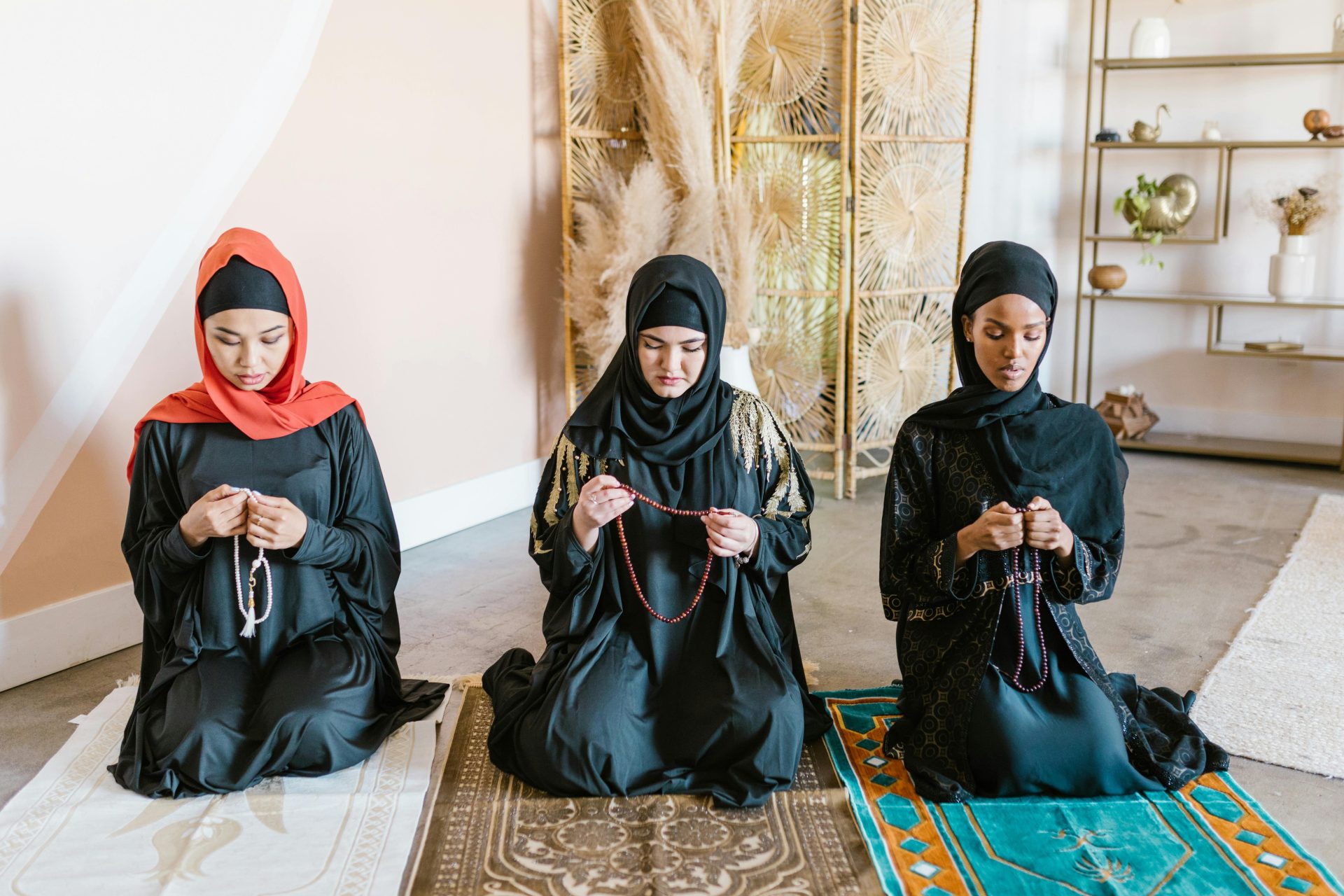 Three women in hijabs pray with beads on rugs indoors, showcasing cultural and religious practices.